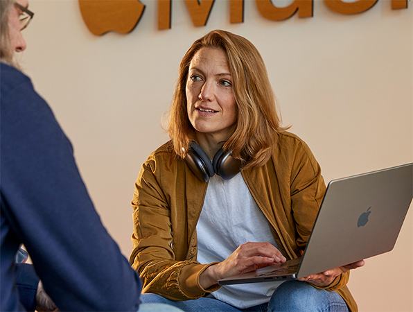 Two Apple Sales and Business Development team members in conversation, one holding a MacBook and sitting in front of a wall with the Apple Music logo on it.