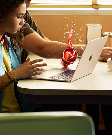 レストランで何かに手を伸ばしている人物が、開いた状態のMacBookの上に冷たい飲み物をこぼしてしまう