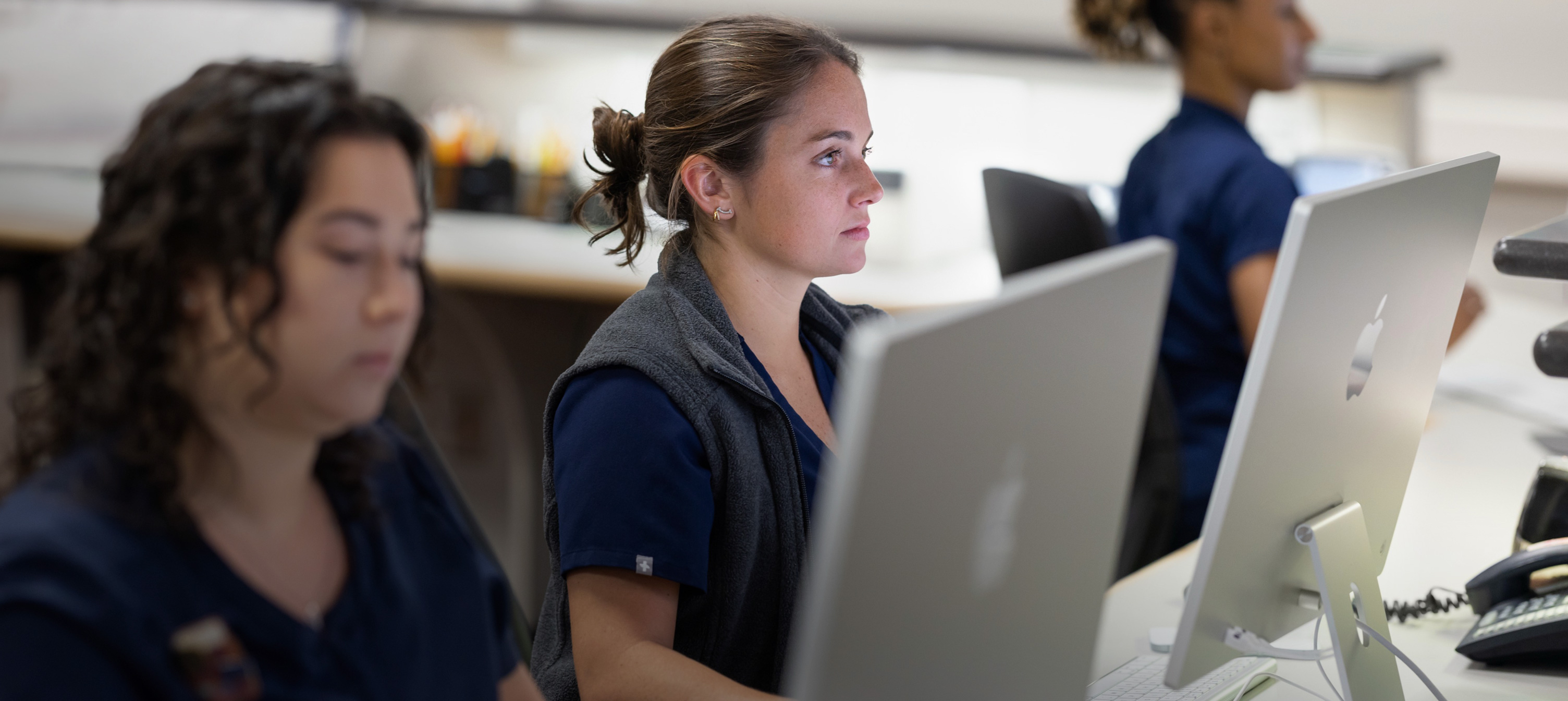 Tres personas de un equipo de enfermería trabajan detrás de pantallas de iMac.
