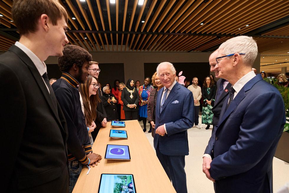 Král Karel a Tim Cook se setkávají s absolventy programu organizace The King's Trust, kteří stojí za stolem s vystavenými iPady.