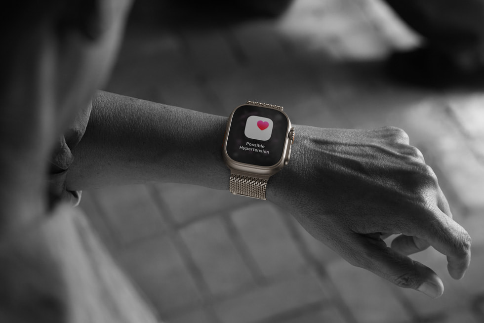 Černobílá fotka osoby, která si na Apple Watch Ultra 3 kontroluje obrazovku oznámení o hypertenzi s ikonou červeného srdce.