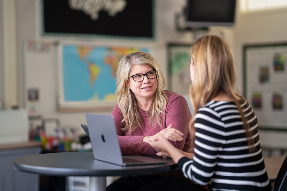 Downey Unified instructional technology coach Marnie Luevano speaks with another adult who is not facing the camera in a classroom setting, with MacBook Air on the table.