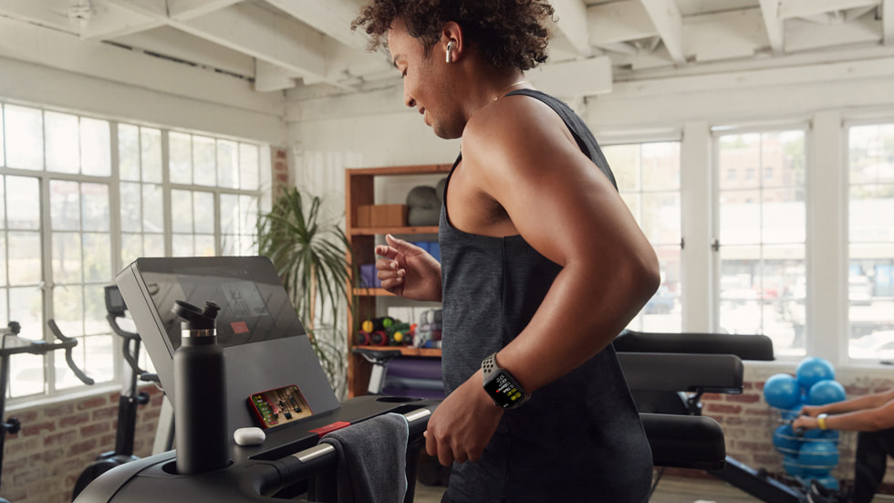 امرأة ترتدي ساعة Apple Watch Series 8 وسماعات AirPods تقوم بتمرين جهاز المشي على Fitness+ باستخدام جهازها iPhone 14.