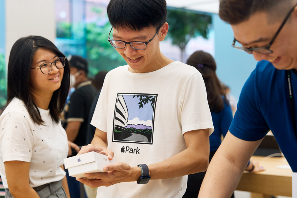عميل في متجر Apple Orchard Road يفتح علبة iPhone 14 بينما تنظر إليه صديقة ويقف أحد أعضاء الفريق مبتسماً.